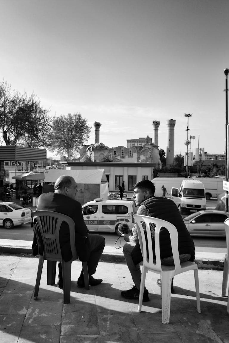 Black and white photo of two men talking, sitting on chairs in a bustling urban area.